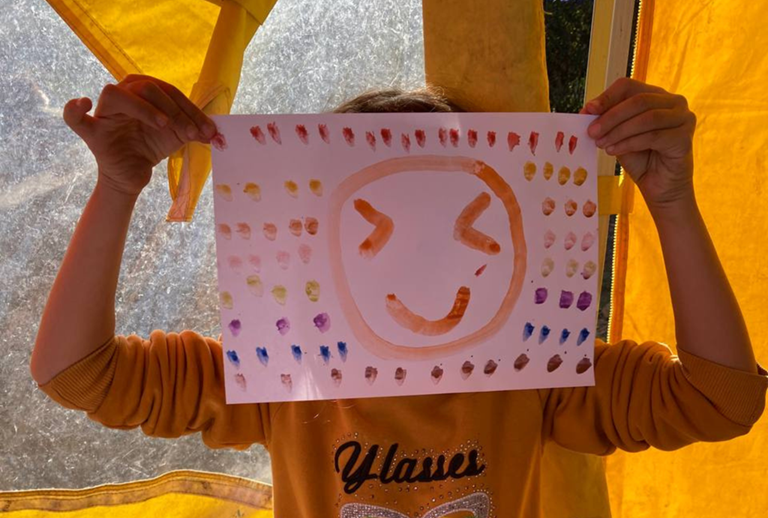 A child holds up a painting in front of thier face. The painting is of a smiley face and the background is decorated with coloured dots. The child is wearing a yellow sweatshirt and is standing inside a yellow tent.